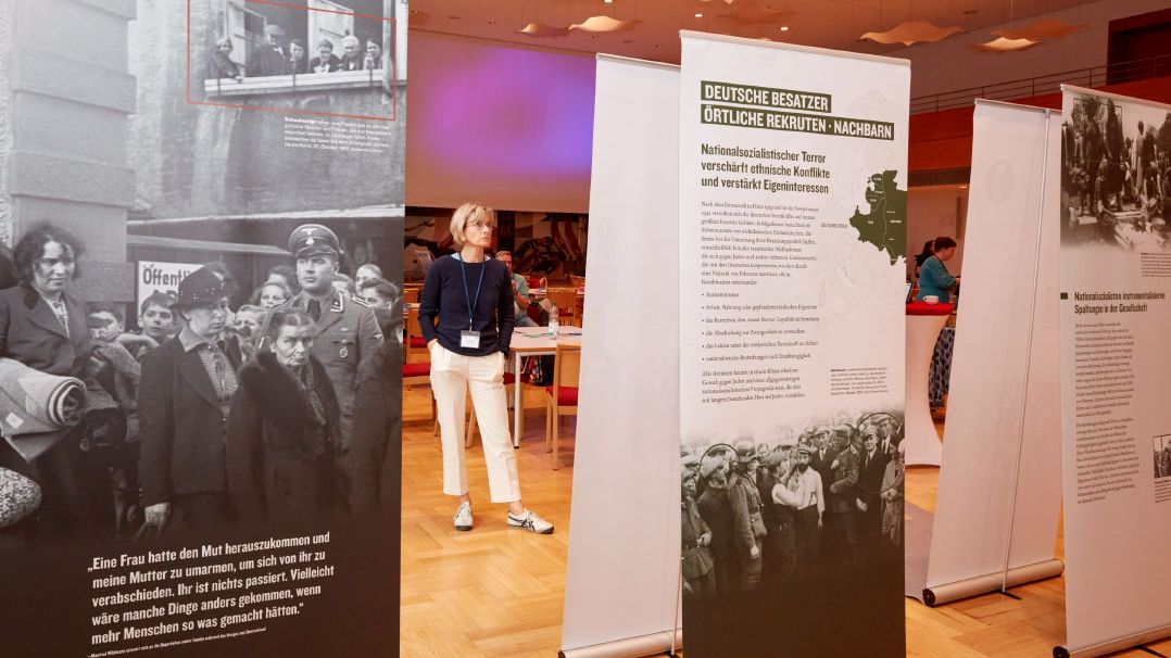 Informationsbanner stehen in einem Raum. Links ein Banner mit dem Text 'Eine Frau hatte den Mut herauszukommen und meine Mutter zu umarmen, im sich von ihr zu verabschieden.' Rechts ein Banner mit dem Titel 'DEUTSCHE BESATZER ÖRTLICHE REKRUTEN - NACHBARN'. Im Hintergrund eine Person in heller Kleidung.