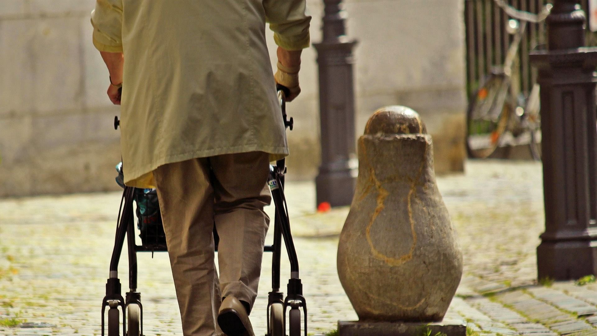 Eine Person in beiger Kleidung läuft mit einem Rollator von der betrachtenden Person weg. Es ist ein gepflasterter Weg, an dem mehrere Poller stehen.