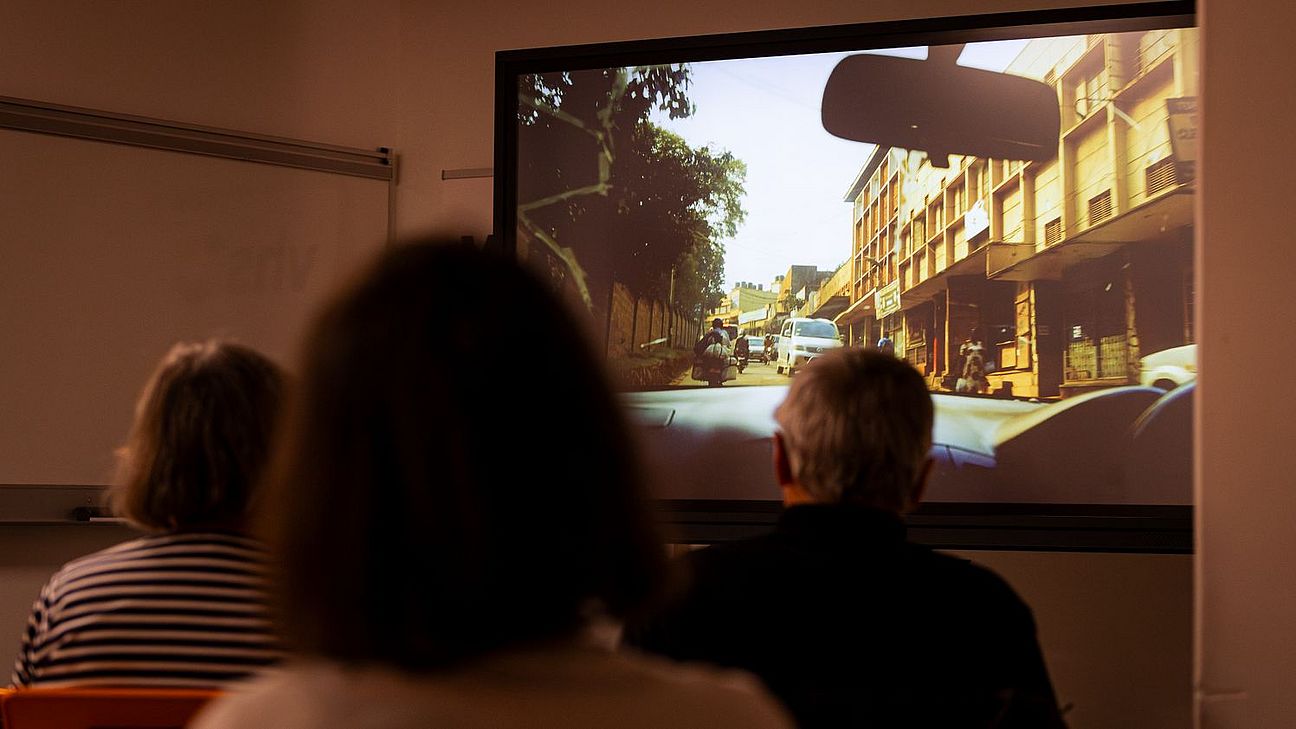 Drei Personen von hinten sitzen in einem dunklen Raum und schauen auf eine Leinwand, die eine Straßenszene mit Autos und Gebäuden zeigt.