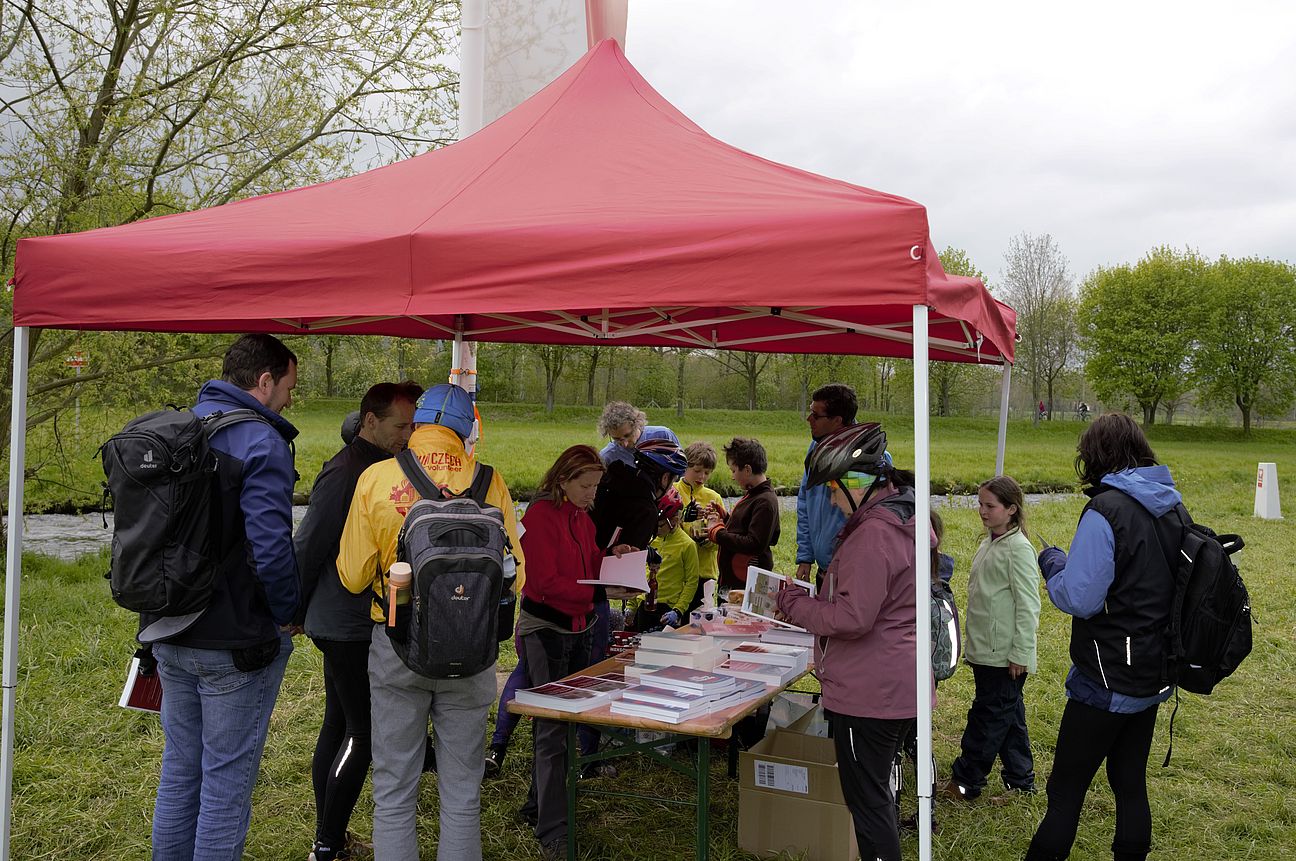 Radtour 8. Mai Ein roter Pavillon, unter dem sich einige Menschen aus der Gruppe die ausgelegten Dinge anschauen.