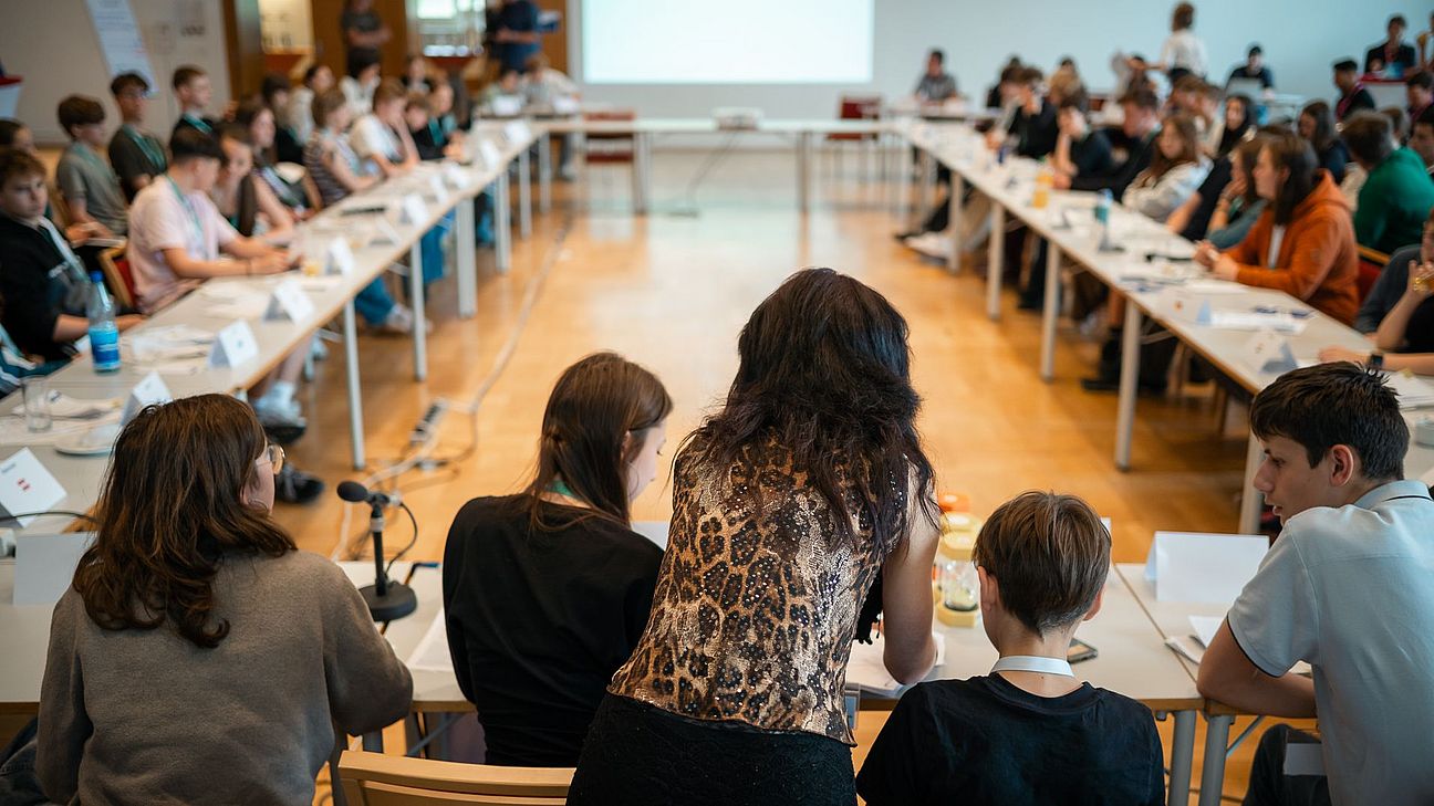 Im Vordergrund beugt sich eine Person über am Tisch sitzende Personen. Im Hintergrund sieht man viele Menschen an Tischen, die U-förmig im Konferenzstil aufgestellt sind.