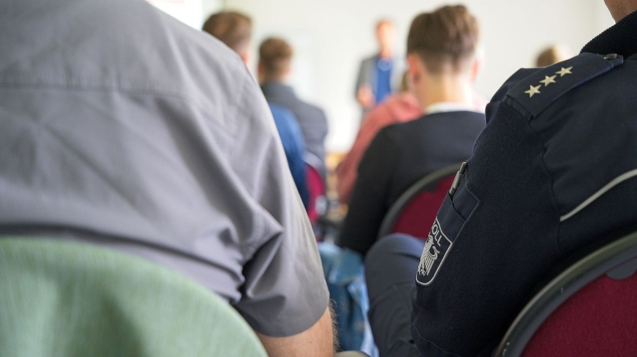 Teilnehmende der Fortbildung sitzen im Seminarraum. Im Vordergrund ist ein Mensch zu sehen, der eine Zolluniform trägt. Im Hintergrund spricht der Referent.