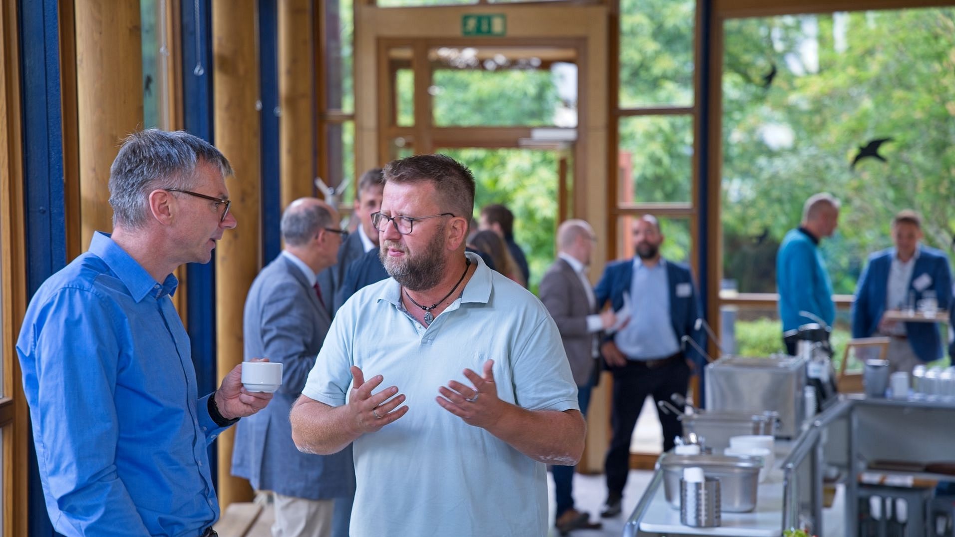 im Vordergrund stehen zwei Personen, die sich unterhalten. Eine hält eine Kaffeetasse, die andere gestikuliert. im Hintergrund sind verschwommen weitere Menschen.