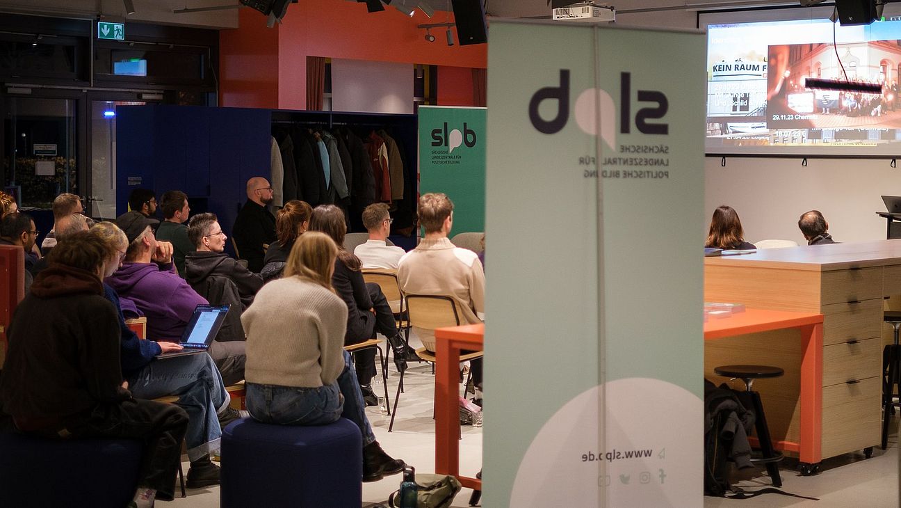 Mehrere Personen sitzen in einem Veranstaltungsraum und schauen auf eine Projektionswand, im Vordergrund ein Roll-up mit dem Logo 'slpb'.