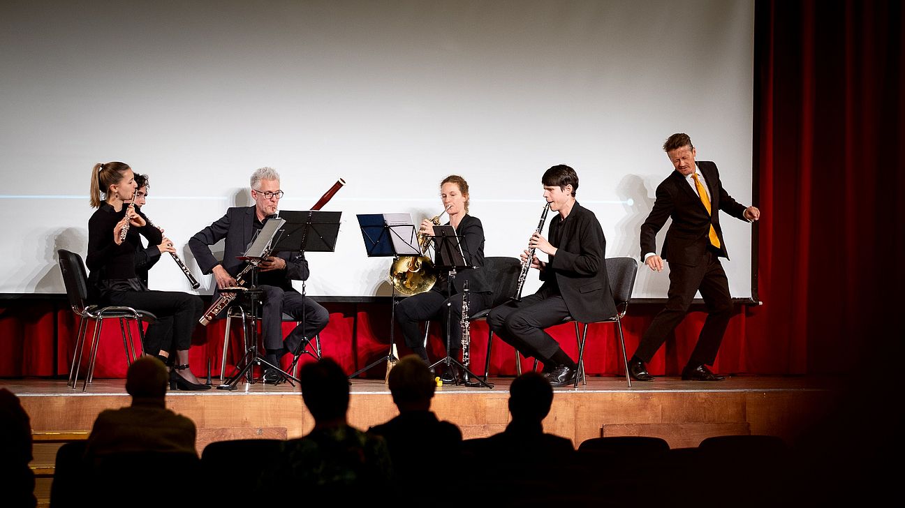 Fünf Musiker auf Stühlen spielen Blasinstrumente auf einer Bühne mit rotem Vorhang, ein Schauspieler steht rechts mit erhobenen Armen.