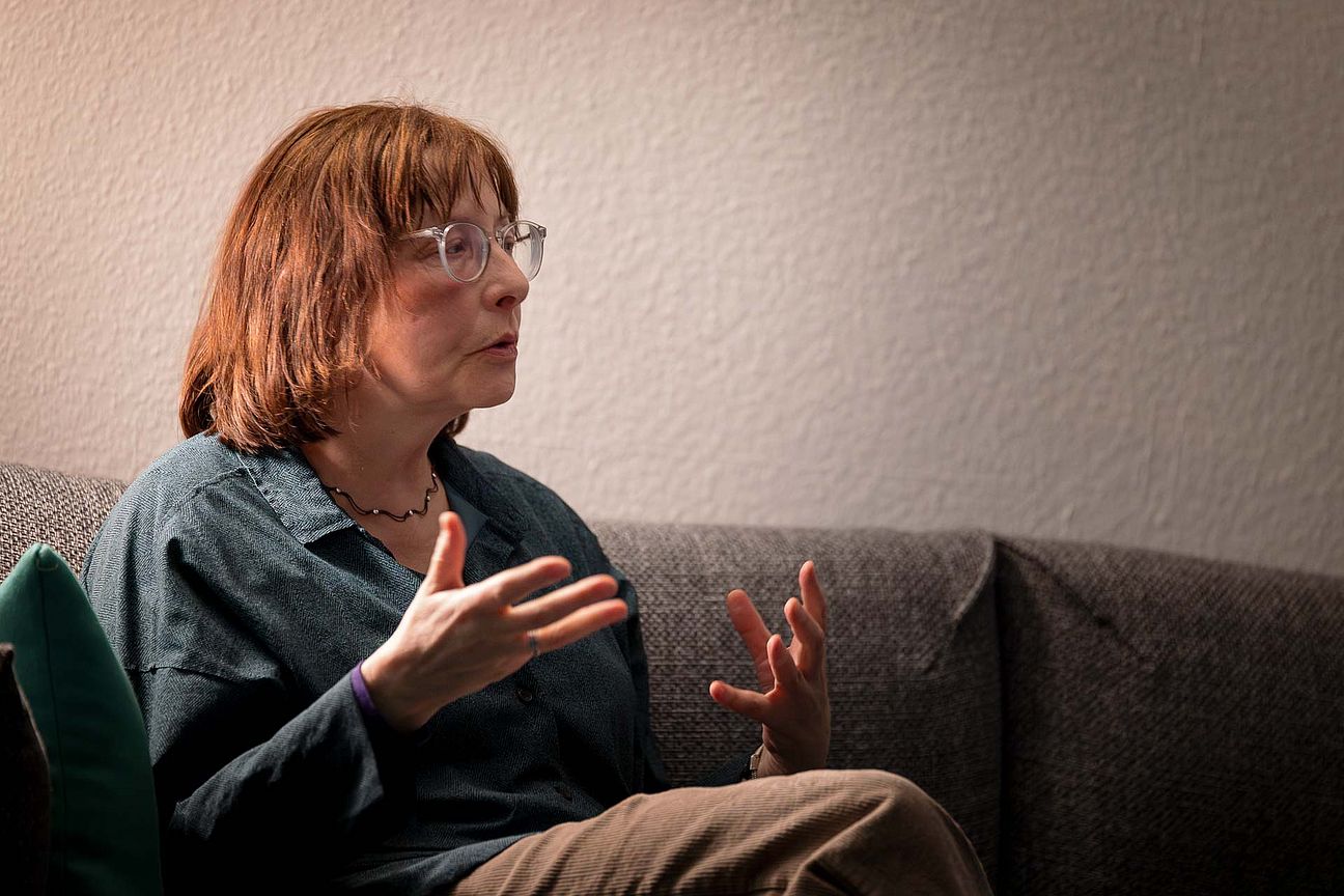 Person mit Brille und braunen Haaren sitzt auf einem Sofa und gestikuliert mit beiden Händen.