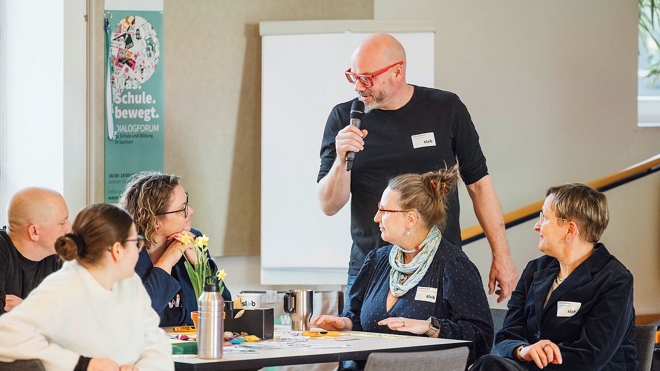KilligBild Eine Person mit Mikrofon steht mittig im Bild an einem tisch mit sitzenden menschen und fragt sie etwas.