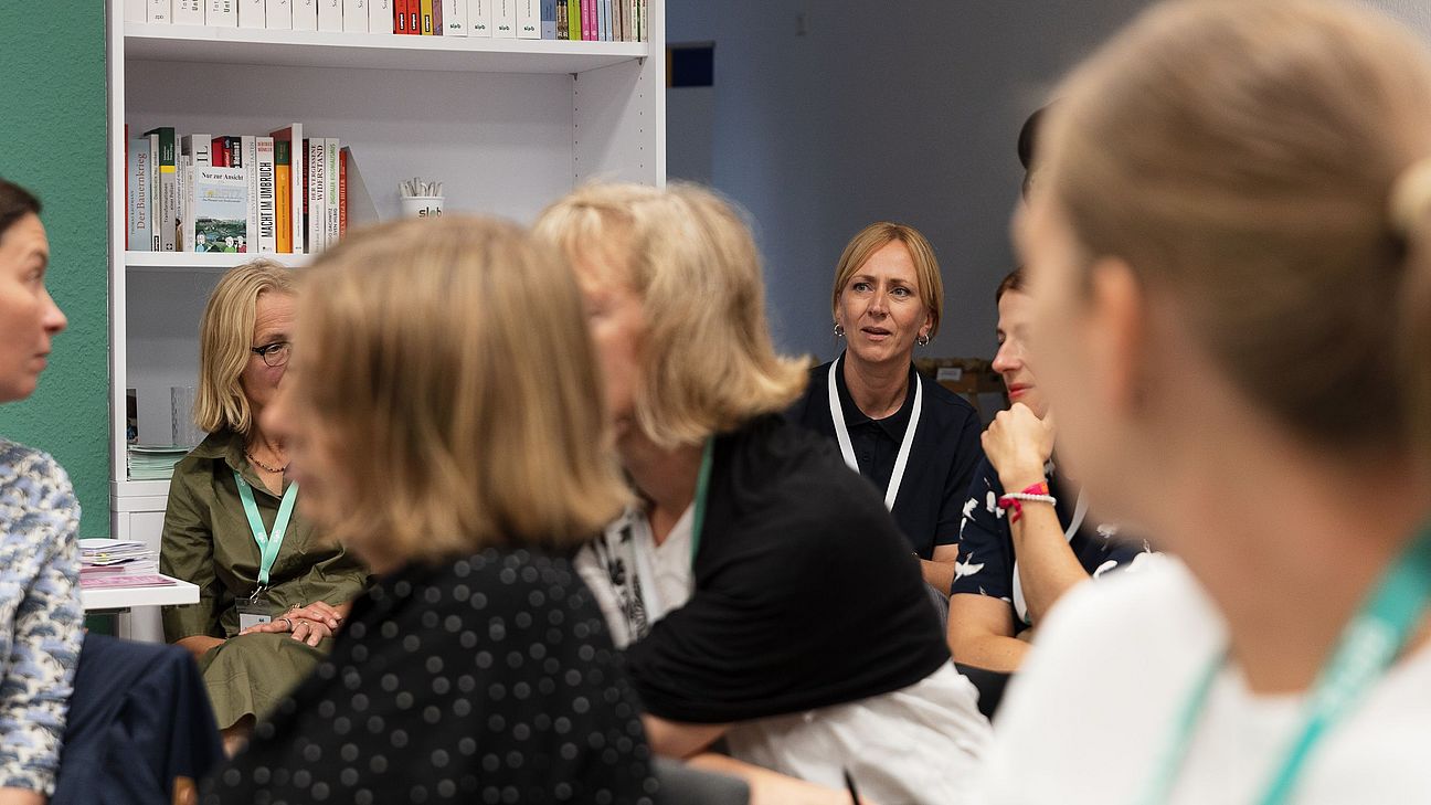 Eine Gruppe von Frauen sitzt in einem Raum. Einige Frauen haben sich umgedreht um sich besser zuhören zu können. Im Hintergrund sind Regale mit Büchern und Ordnern sichtbar.