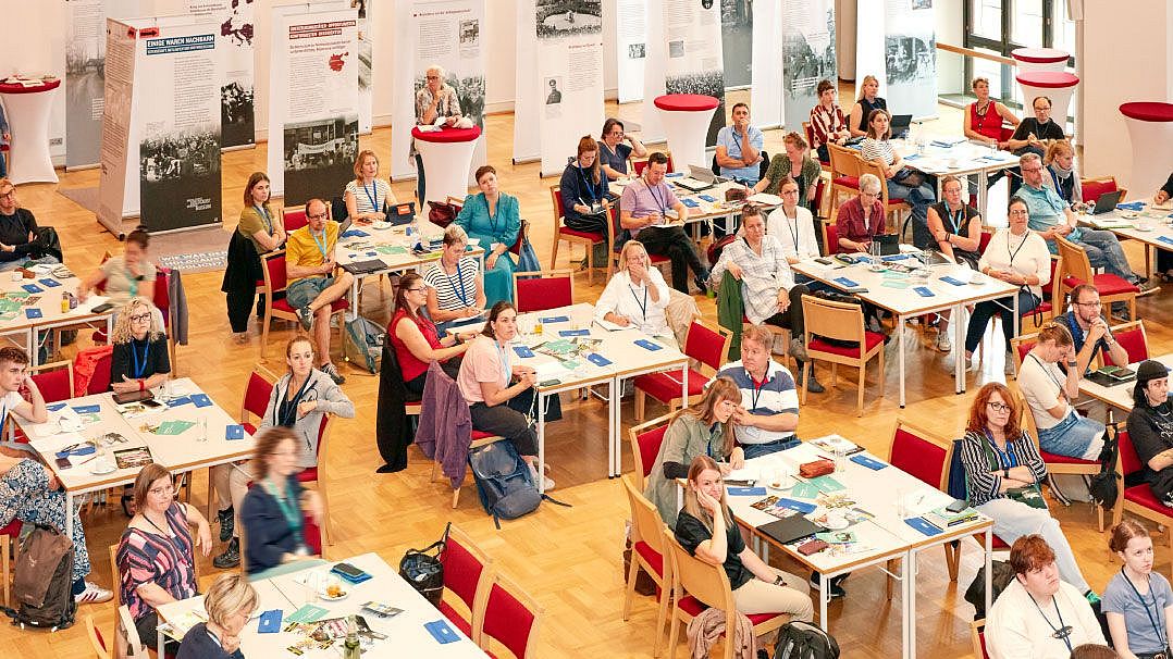 Eine große Gruppe von Menschen sitzt an Tischen in einem Raum. Im Hintergrund sind Informationsplakate aufgestellt. Einige Personen haben Notizen und Materialien vor sich.