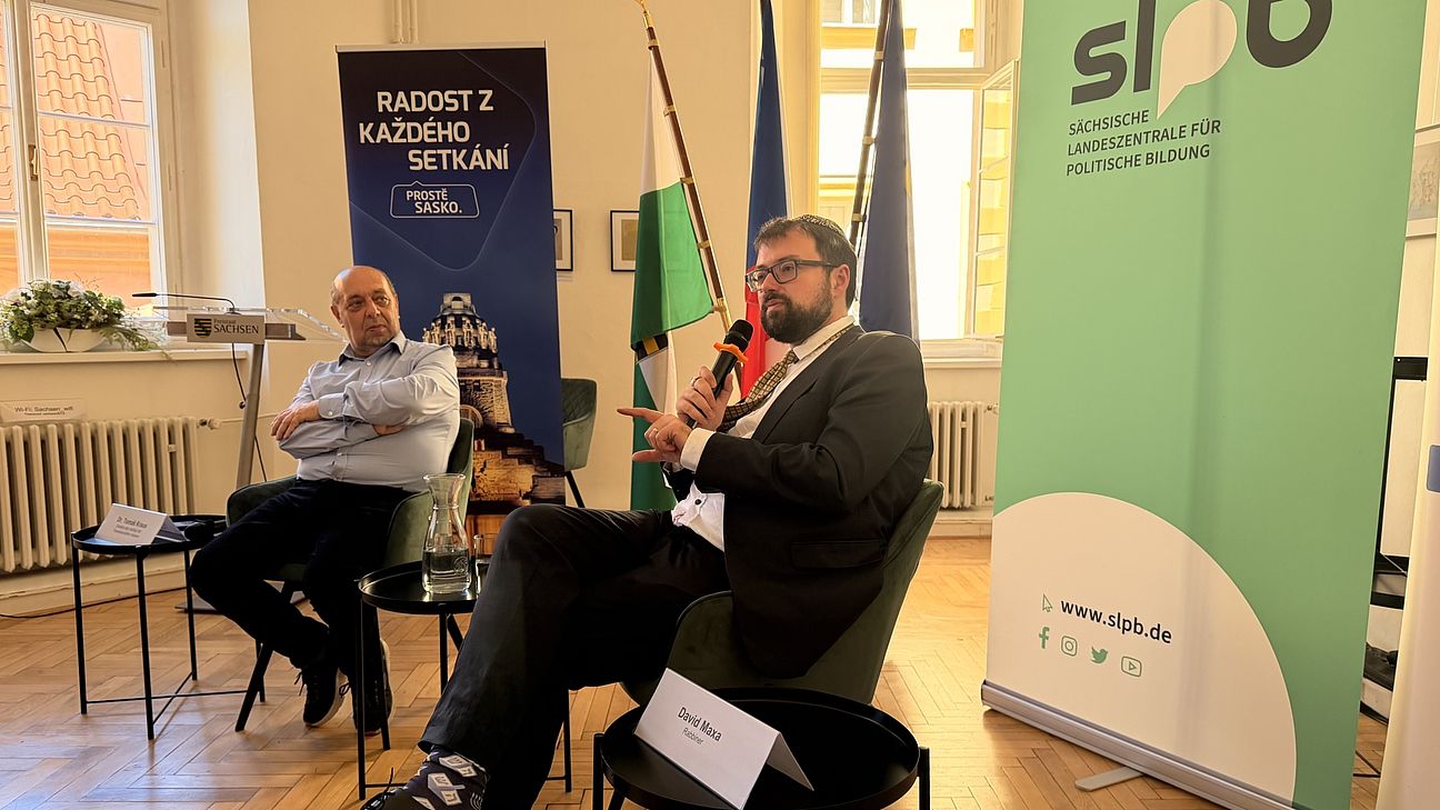 Zwei Männer sitzen auf Stühlen, einer hält ein Mikrofon. Im Hintergrund sind Banner zu sehen. Eins von der SLpB. dazu drei Flaggen von Sachsen, Tschechien und der EU.