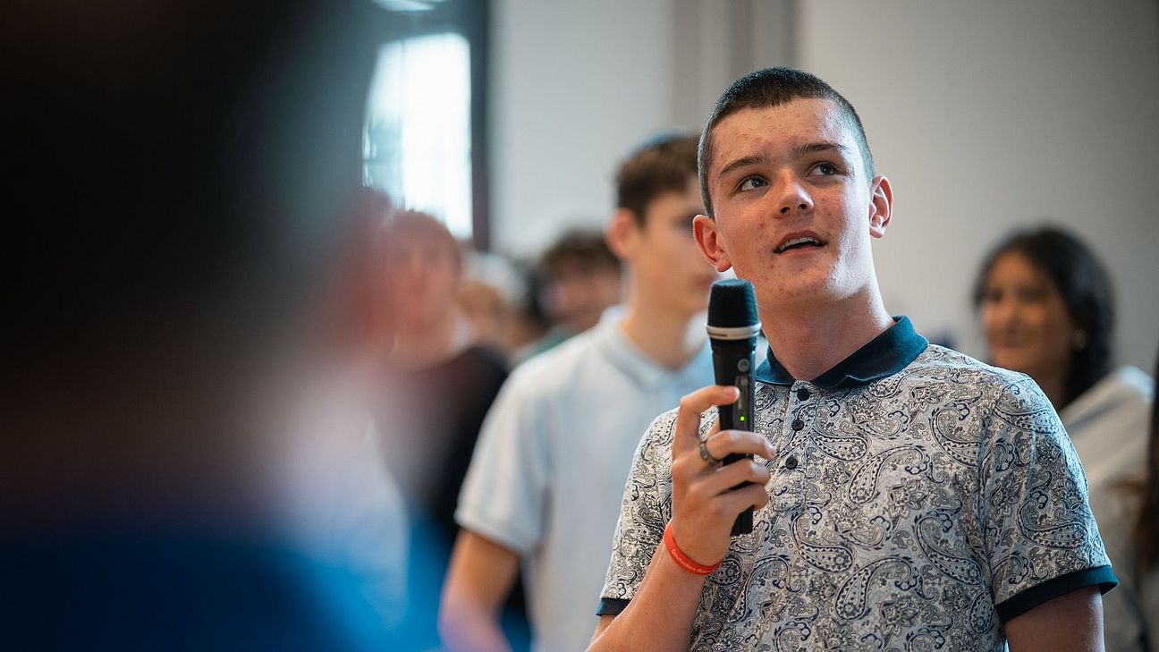 Scharf im Vordergrund steht eine junge Person mit Mikrofon, dahinter weitere Personen unscharf.