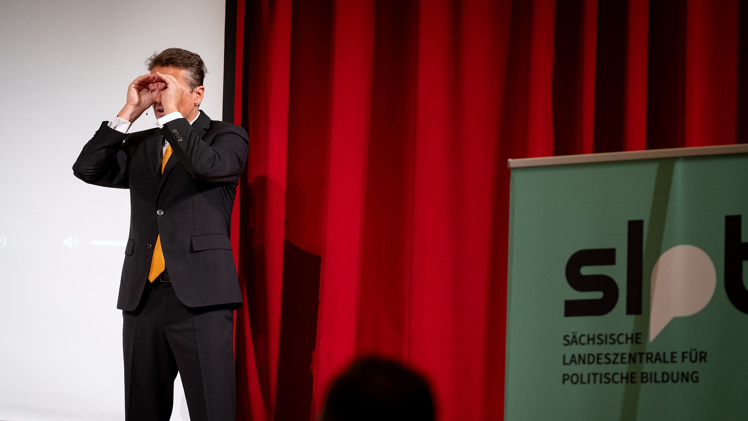 Mann im schwarzen Anzug mit orangefarbener Krawatte steht vor rotem Vorhang und weißer Leinwand, rechts ein Banner mit Schriftzug 'slpb Sächsische Landeszentrale für politische Bildung'.