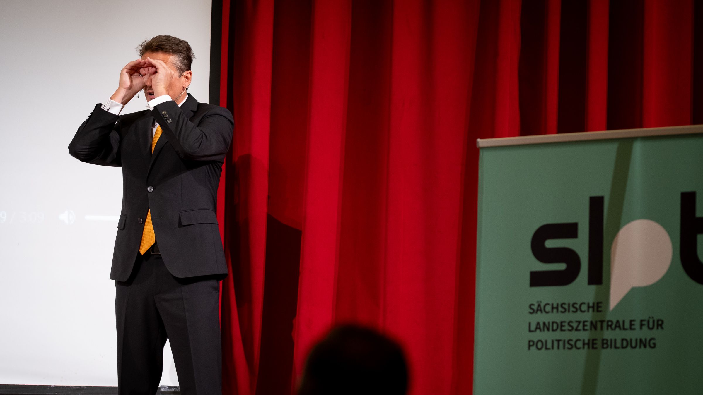 Mann im schwarzen Anzug mit orangefarbener Krawatte steht vor rotem Vorhang und weißer Leinwand, rechts ein Banner mit Schriftzug 'slpb Sächsische Landeszentrale für politische Bildung'.