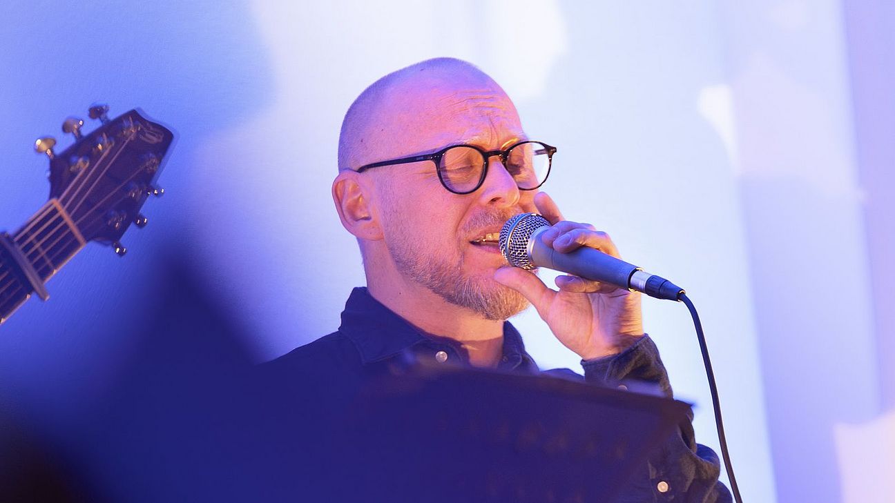 Dr. Christoph Meißelbach singt mit geschlossenen Augen in ein Handmikrofon. Er trägt eine dunkle Jacke und eine runde Brille. Die Bühne ist in blau-violettes Licht getaucht.