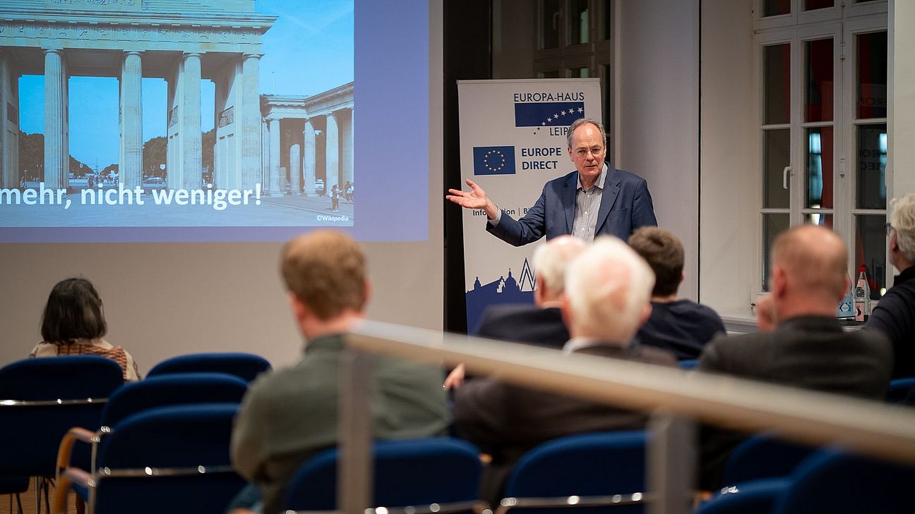 Mann steht vor Publikum und hält Vortrag mit Projektion des Brandenburger Tores und Text '... mehr, nicht weniger!'