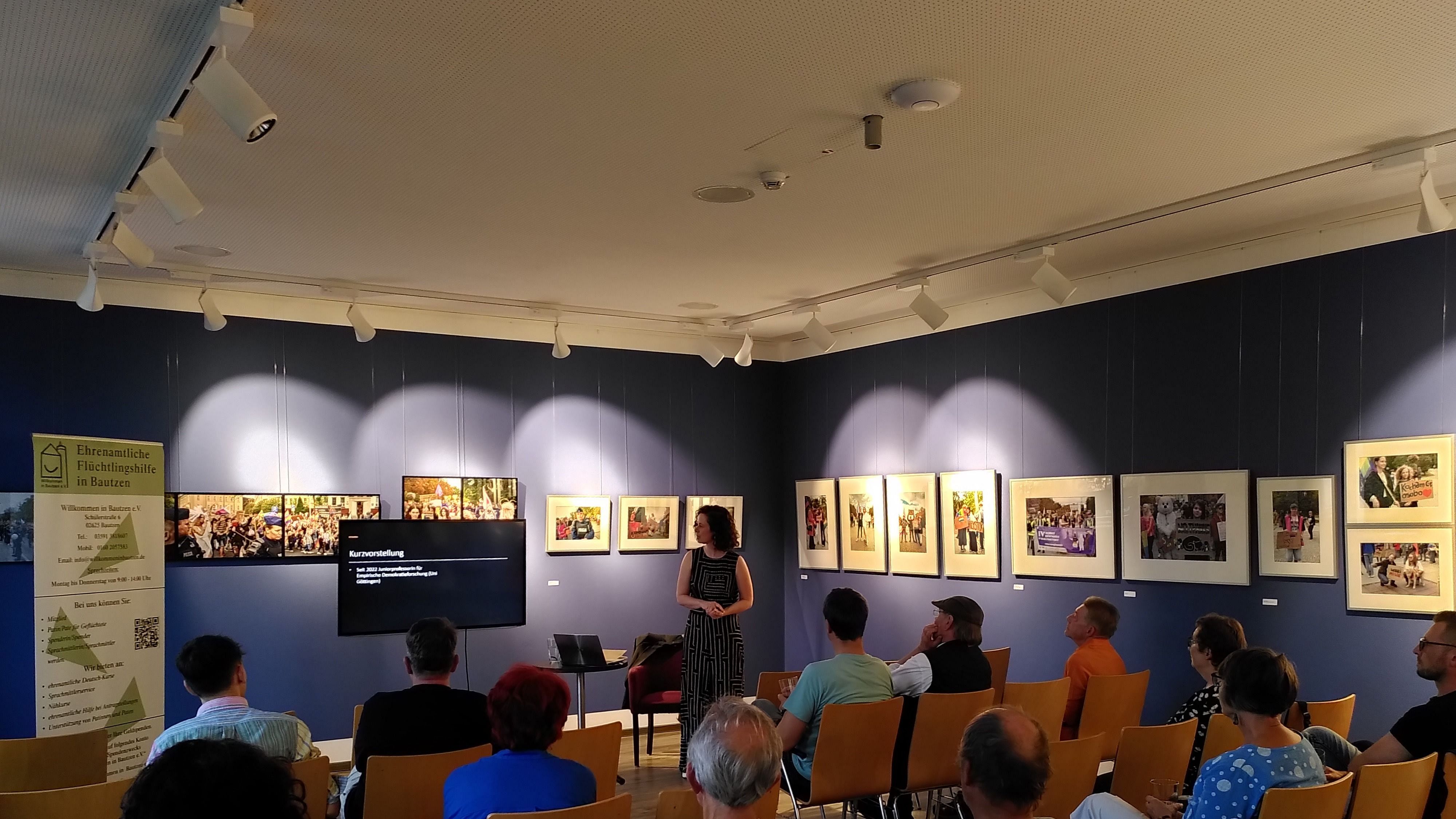 JProf. Dr. Theres Matthieß steht vor einer Präsentation in einem Raum mit Publikum. An der Wand hängen Bilder. Stühle sind in Reihen angeordnet.