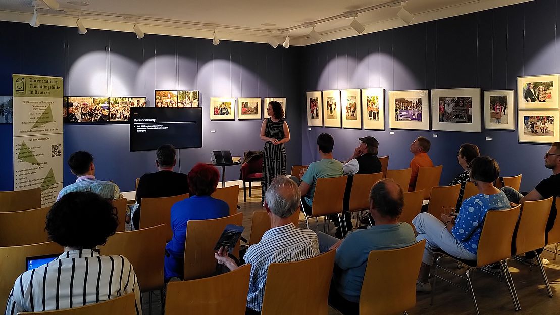 JProf. Dr. Theres Matthieß steht vor einer Präsentation in einem Raum mit Publikum. An der Wand hängen Bilder. Stühle sind in Reihen angeordnet.