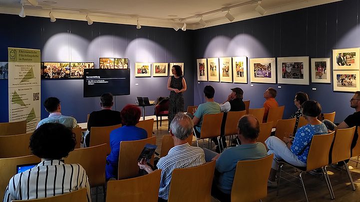 JProf. Dr. Theres Matthieß steht vor einer Präsentation in einem Raum mit Publikum. An der Wand hängen Bilder. Stühle sind in Reihen angeordnet.
