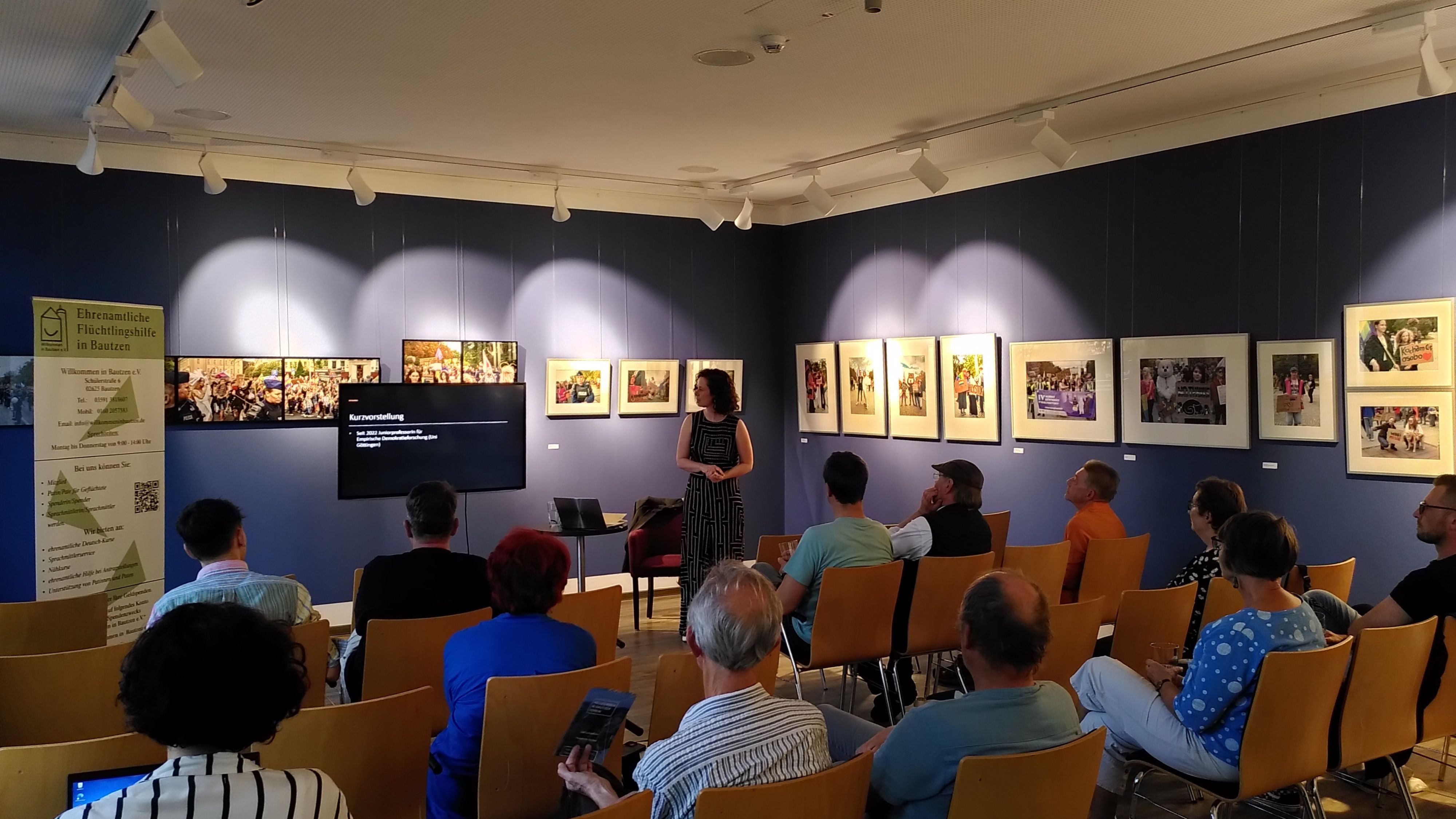 JProf. Dr. Theres Matthieß steht vor einer Präsentation in einem Raum mit Publikum. An der Wand hängen Bilder. Stühle sind in Reihen angeordnet.