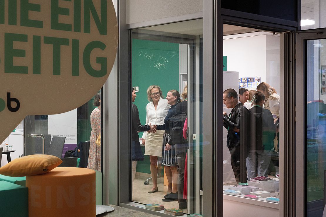 Blick durch Fenster: Gruppierung von Personen in einem Raum, einige stehen. Sichtbare Möbel und Bücher auf einem Tisch.