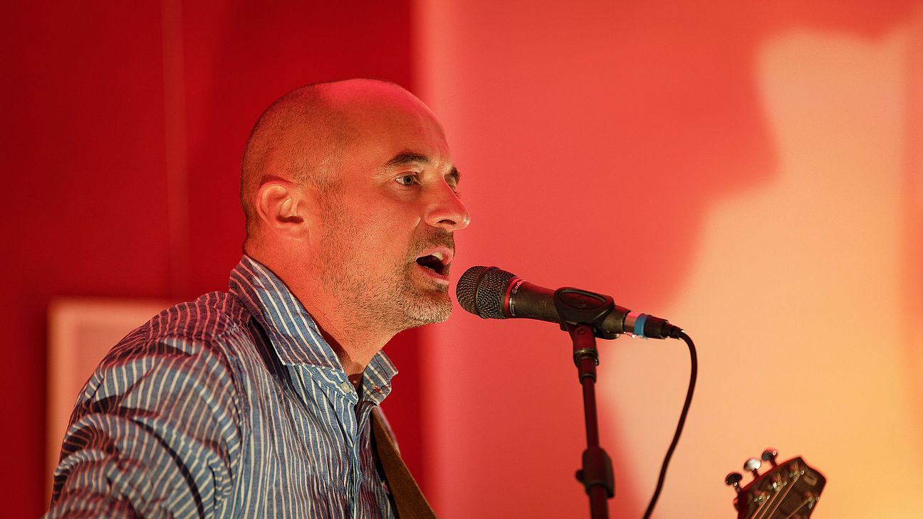 Christoph Schlosser singt in ein Mikrofon. Er trägt ein blau gestreiftes Hemd und einen Gitarrengurt über der Schulter. Im Hintergrund ist die Bühne in rotes Licht getaucht.