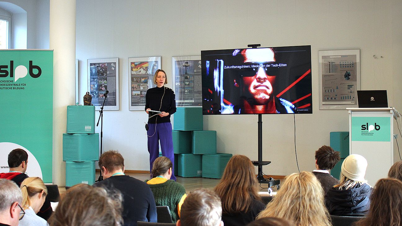 Mehrere Personen sitzen in einem Raum mit weißer Wand, schauen auf einen großen Bildschirm auf einem Ständer. Links steht ein grüner Banner mit dem Schriftzug 'slpb'. Rechts steht ein weißes Podium mit dem gleichen Logo und einem Laptop. Im Hintergrund sind grüne Kisten und gerahmte Bilder an der Wand zu sehen.
