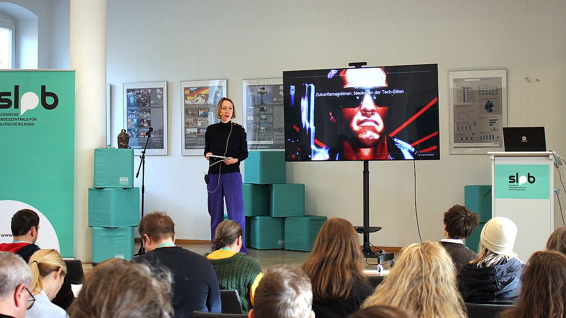 Mehrere Personen sitzen in einem Raum mit weißer Wand, schauen auf einen großen Bildschirm auf einem Ständer. Links steht ein grüner Banner mit dem Schriftzug 'slpb'. Rechts steht ein weißes Podium mit dem gleichen Logo und einem Laptop. Im Hintergrund sind grüne Kisten und gerahmte Bilder an der Wand zu sehen.