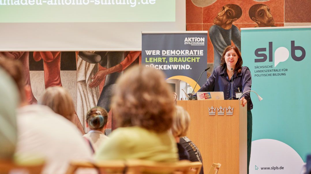 Eine Person steht an einem Rednerpult mit einem Logo der 'Sächsischen Landeszentrale für politische Bildung'. Im Hintergrund sind Banner mit Texten zu Demokratie und einer künstlerischen Wandmalerei zu sehen.
