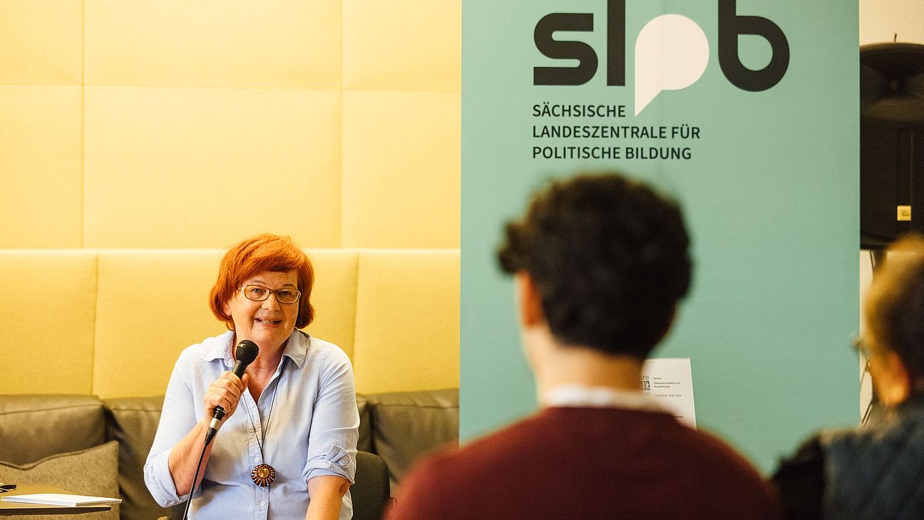 Frau mit Mikrofon sitzt auf einem Stuhl vor einem Banner der Sächsischen Landeszentrale für politische Bildung, zwei Personen im Vordergrund von hinten zu sehen.