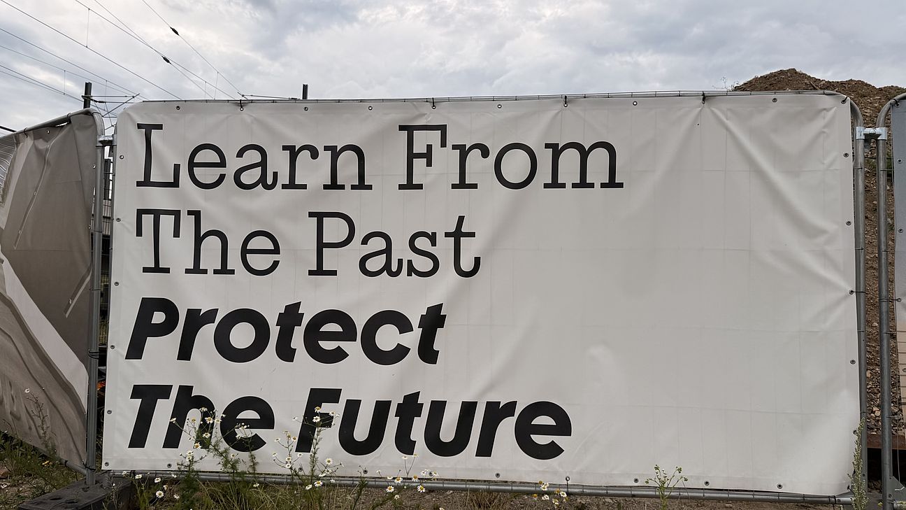 An einem Baustellenzaun hängt ein Banner mit der Aufschrift "learn from the past, protect the future"