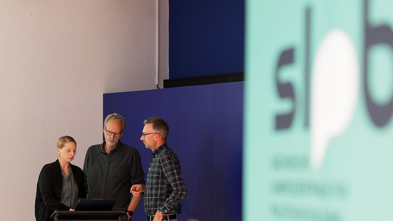 Drei Personen stehen bei einer Veranstaltung an einem Rednerpult und besprechen sich vor einer Präsentation. Im Vordergrund ist unscharf das Logo der Sächsischen Landeszentrale für politische Bildung zu sehen.