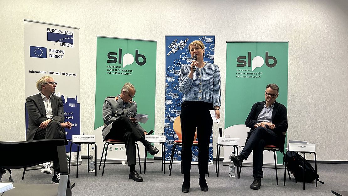 Vor Roll Ups sitzen drei Menschen. Zwischen ihnen steht Jana Puglierin, Leiterin des ECFR mit einem Mikrofon in der Hand