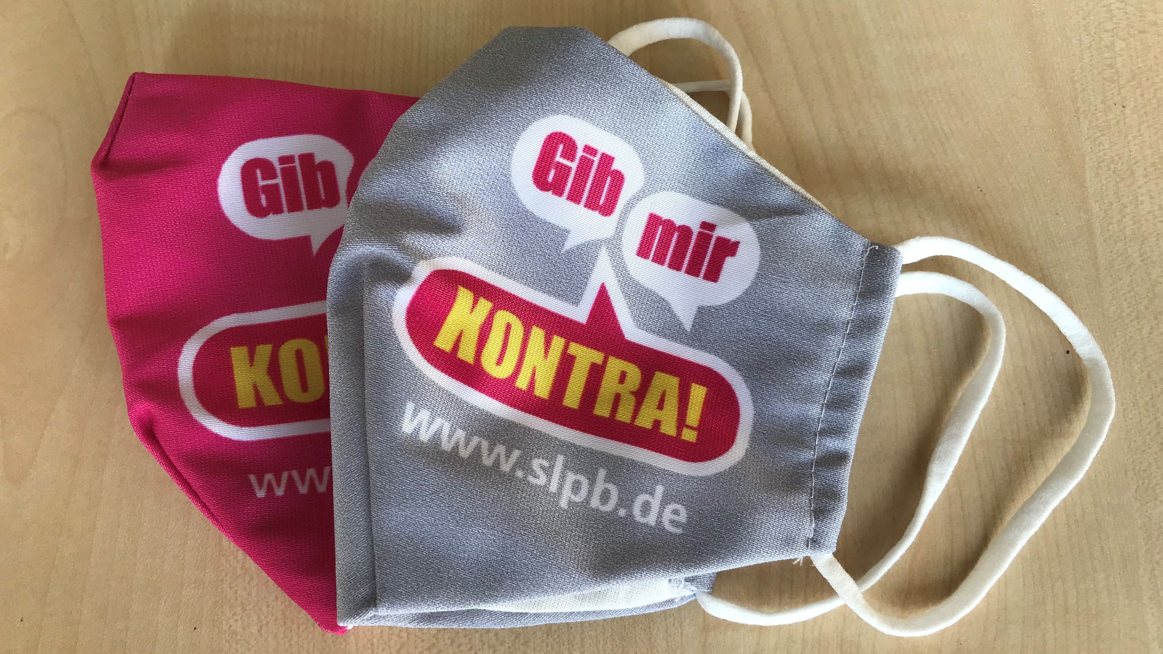 Gib mir Kontra Maske Zu sehen sind zwei Gesichtsmasken, eine graue und eine pinke, die auf einer hellen Holzoberfläche liegen. Beide Masken haben den gleichen Aufdruck: zwei weiße Sprechblasen mit rotem Text "Gib mir" und eine gelbe Sprechblase mit rotem Text "KONTRA!". Darunter ist die Webseite "www.slpb.de" in Weiß gedruckt. Bei Klick vergrößert sich das Bild.