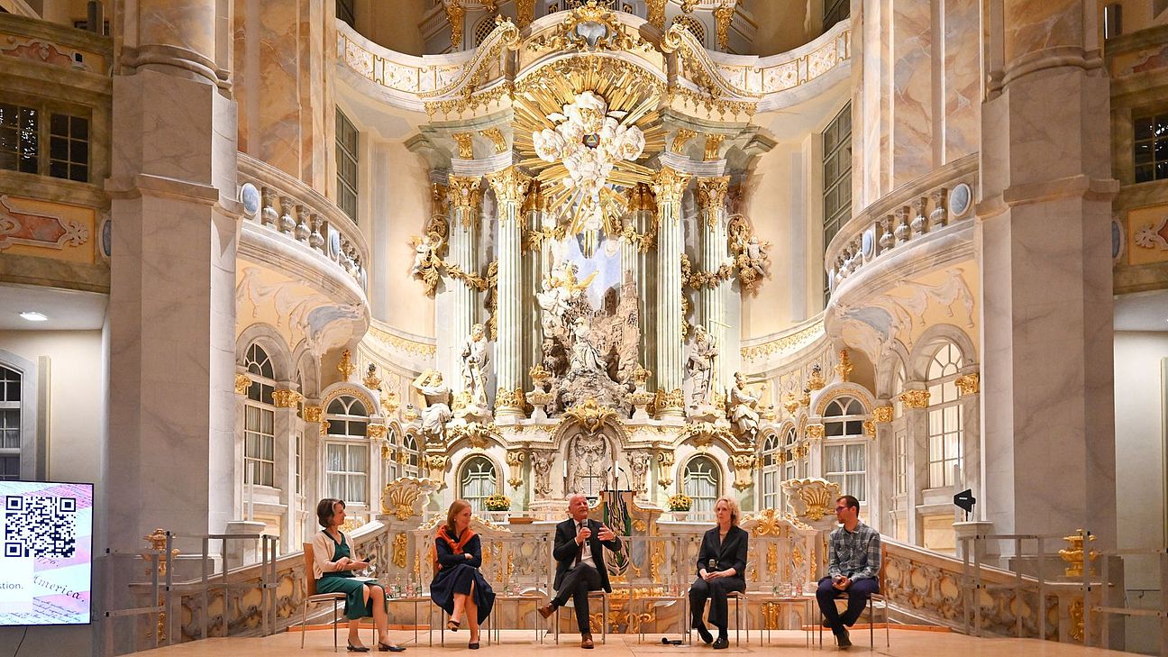 Fünf Personen sitzen auf Stühlen vor einer barocken Altarwand in der Frauenkirche Dresden.