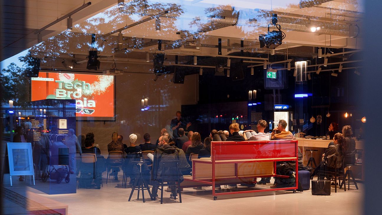 Durch eine Glasfront ist ein Veranstaltungsraum mit Publikum zu sehen. Auf der Leinwand steht „Tech Bro Utopia“, während Menschen einem Vortrag folgen.