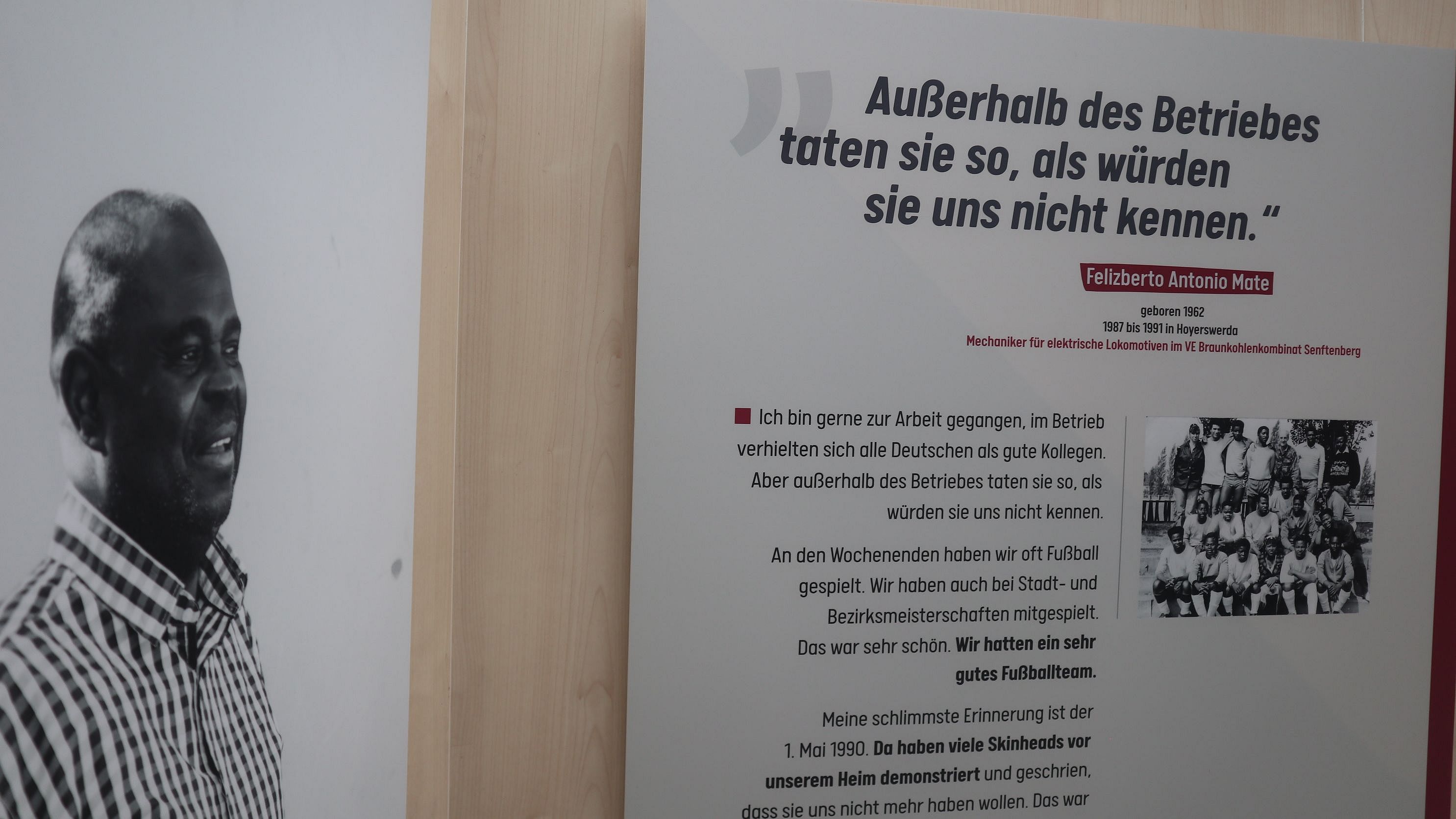 Ausstellung in Hoyerswerda Das Bild zeigt einen Ausstellungsbereich mit einem großen Zitat an der Wand. Das Zitat lautet: "Außerhalb des Betriebes taten sie so, als würden sie uns nicht kennen." und wird Teitótebo Antonio Mate zugeschrieben, einem Zeitzeugen aus Mozambique. Neben dem Zitat befindet sich ein kleinerer Text, der mit "Ich bin gerne zur Arbeit gegangen, im Betrieb verhielten sich alle Deutschen ganz kollegial. Aber außerhalb des Betriebes taten sie so, als würden sie uns nicht kennen." beginnt. Es gibt eine weitere Erläuterung seiner Erfahrungen und Erinnerungen, die mit einem Fußballspiel und einer persönlichen Erinnerung an den 1. Mai 1980 endet. Auf der linken Seite ist ein Porträt von Antonio Mate zu sehen. Bei Klick vergrößert sich das Bild.