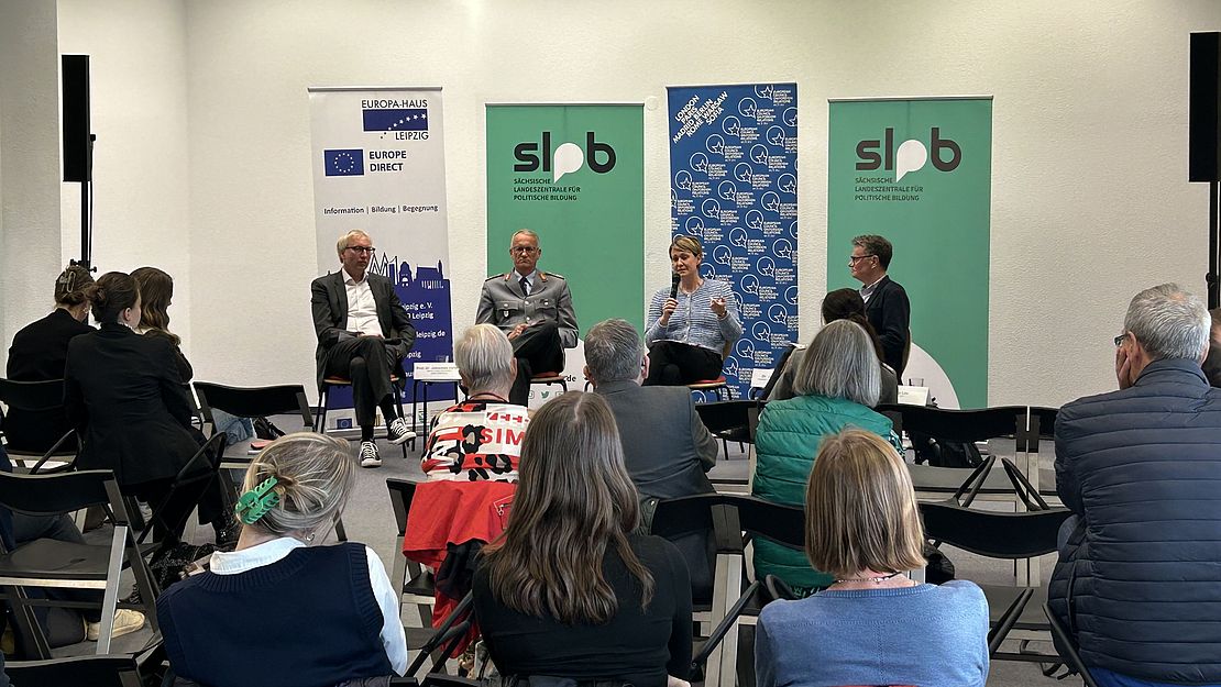 Im Vordergrund sind die Teilnehmerinnen der Veranstaltung auf ihren Stühlen sitzend zu sehen. Im Hintergrund sitzt das Podium.