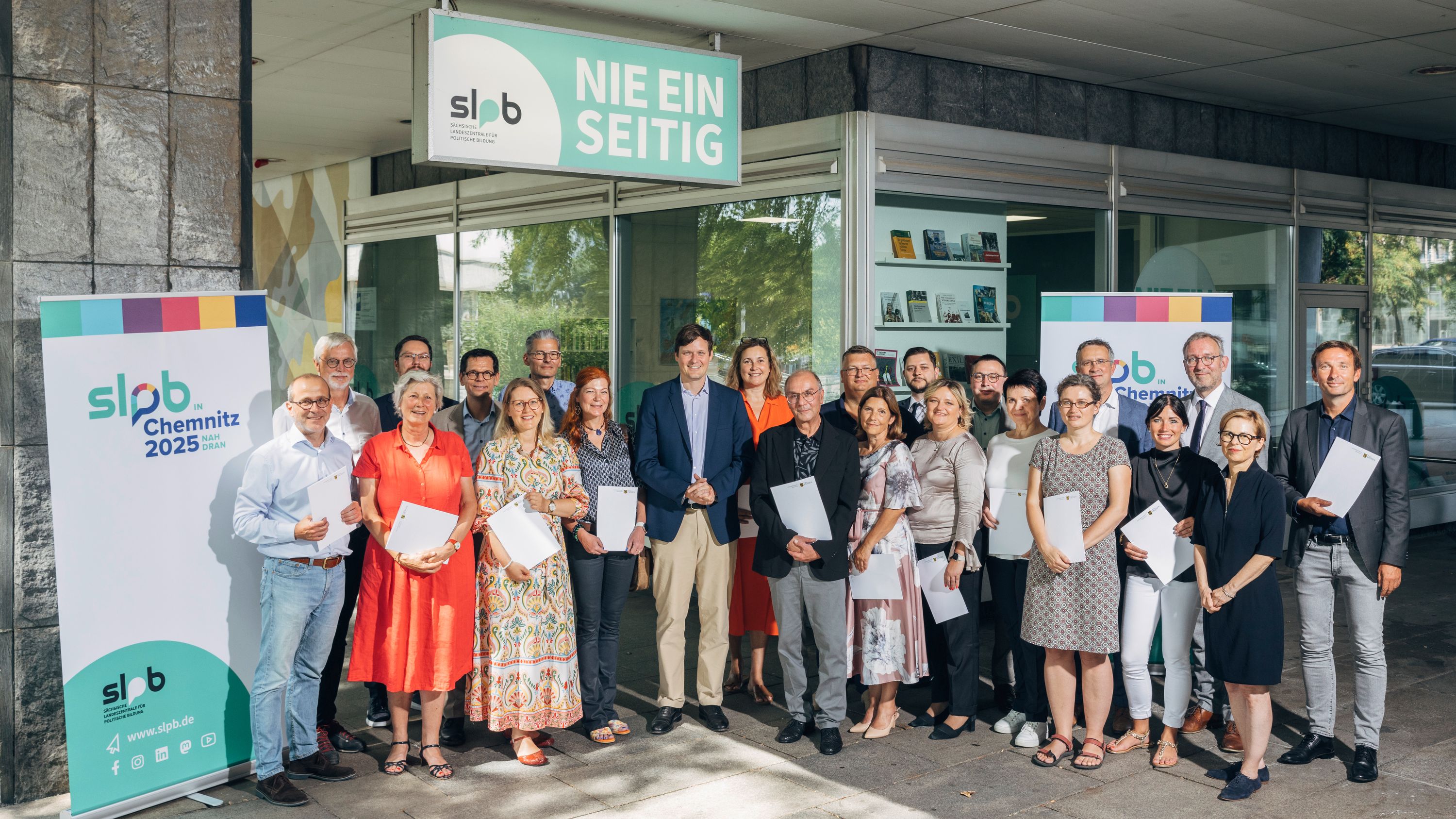 Gruppenfoto des Kuratoriums der Sächsischen Landeszentrale für politische Bildung vor dem Gebäude mit Bannern 'slpb Chemnitz 2025' und 'Nie einseitig'