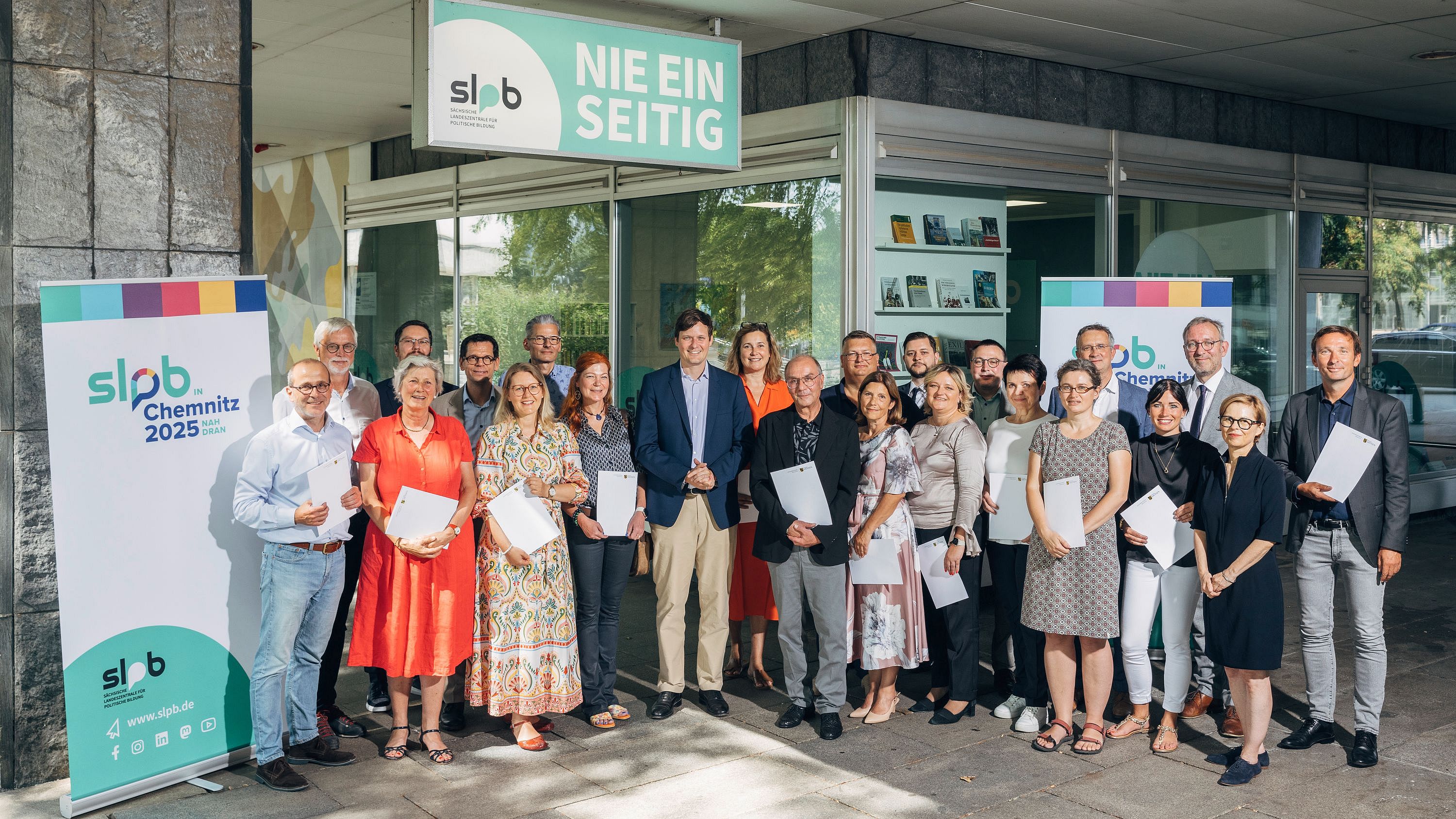 Gruppenfoto des Kuratoriums der Sächsischen Landeszentrale für politische Bildung vor dem Gebäude mit Bannern 'slpb Chemnitz 2025' und 'Nie einseitig'