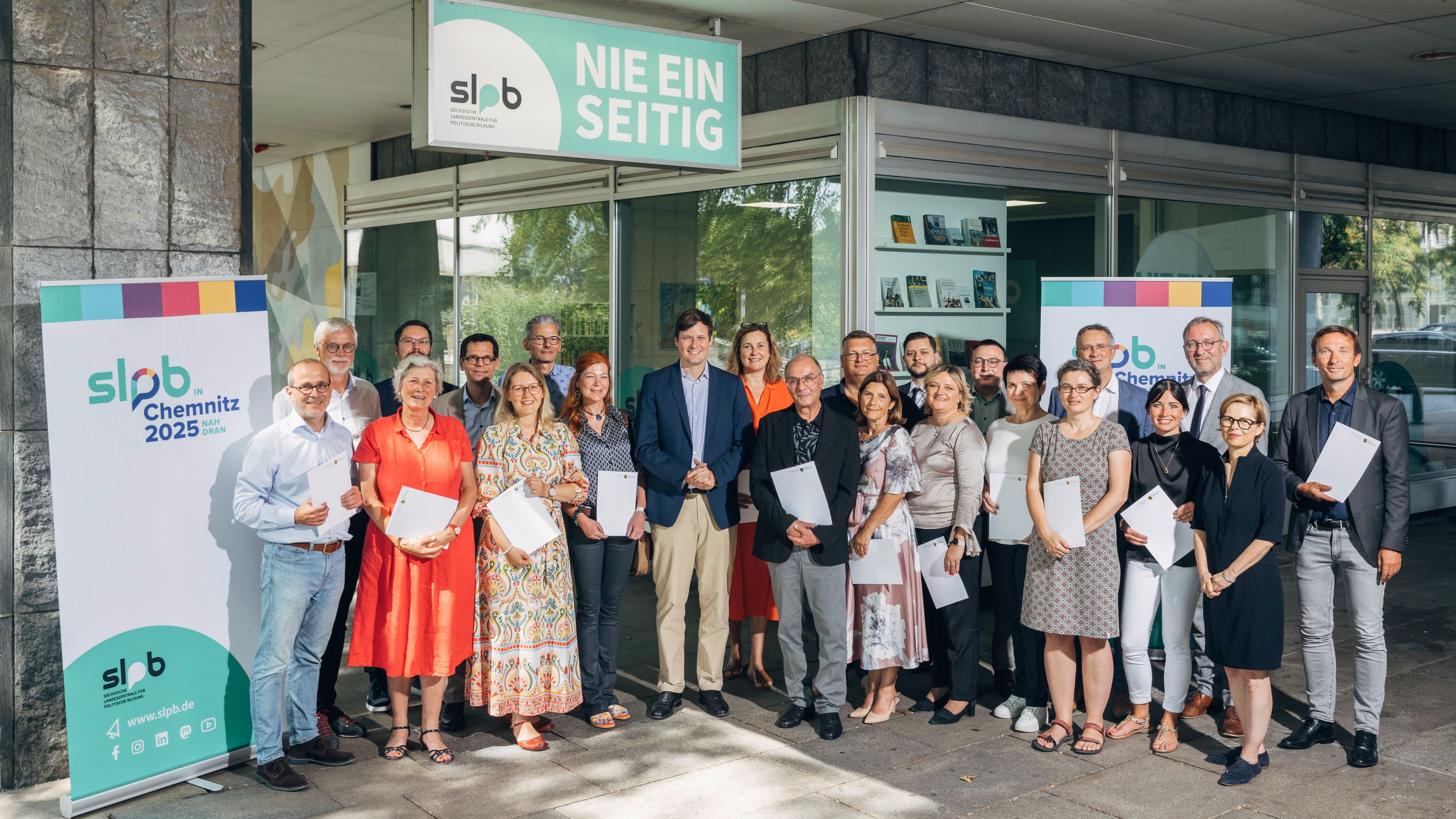 Gruppenfoto des Kuratoriums der Sächsischen Landeszentrale für politische Bildung vor dem Gebäude mit Bannern 'slpb Chemnitz 2025' und 'Nie einseitig'