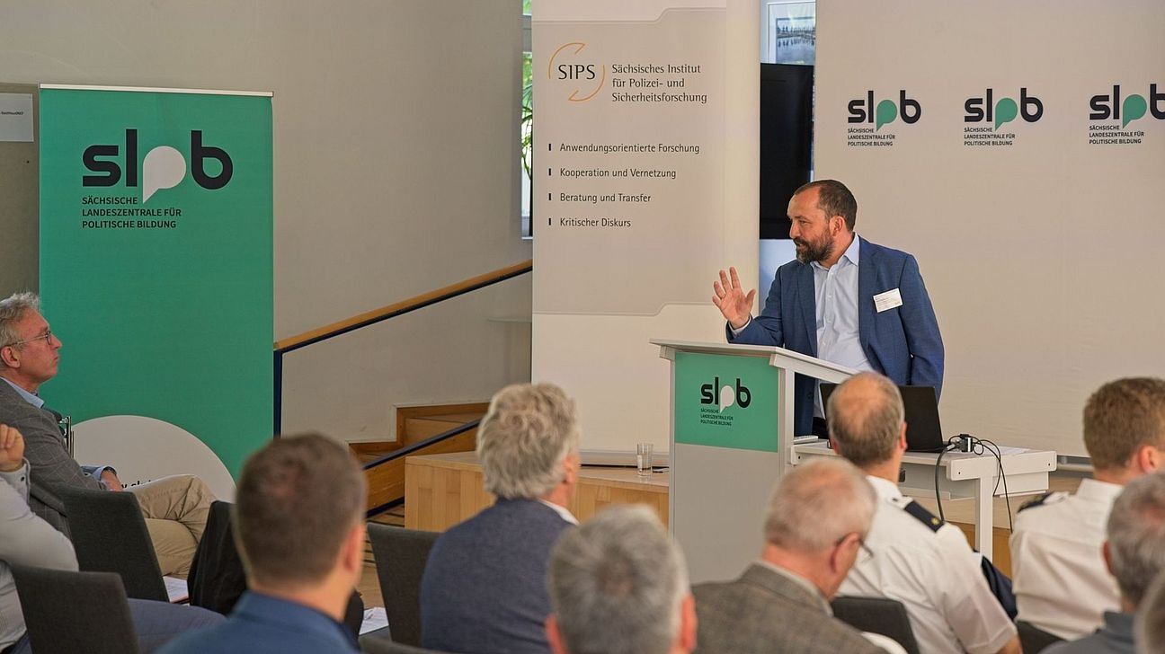 Eine Person steht an einem Pult, spricht und gestikuliert. im Vordergrund sitzen Menschen, der redenden Person zugewandt. Im Hintergrund Banner und Wände der Kooperationspartner.