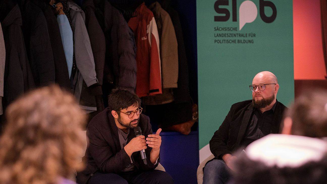 Zwei Männer sitzen auf Stühlen, einer spricht in ein Mikrofon, im Hintergrund hängen mehrere Jacken an einer Garderobe, rechts ein Banner mit dem Logo der Sächsischen Landeszentrale für politische Bildung.