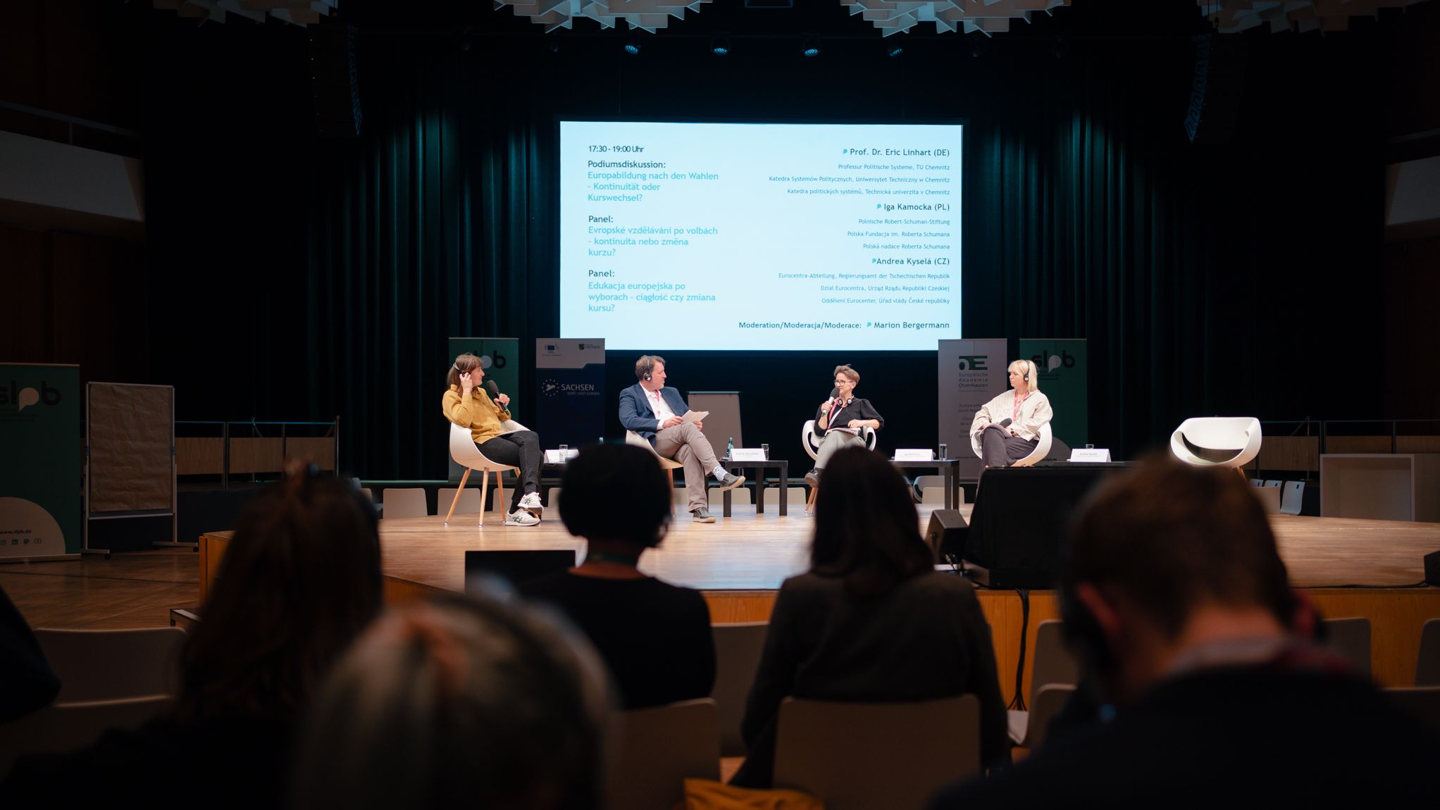 Podium mit vier Personen, drei Frauen und ein Mann, sitzen auf Stühlen auf einer Bühne vor einer großen Leinwand mit Text, Publikum im Vordergrund sichtbar.
