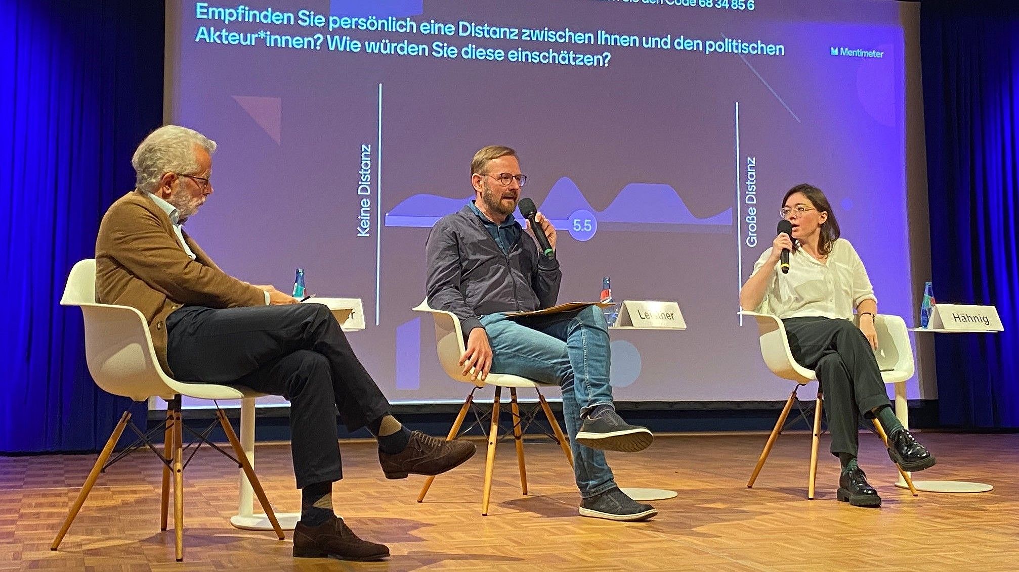 Das Bild zeigt eine Podiumsdiskussion mit drei Personen, die auf Stühlen auf einer Bühne sitzen. Im Hintergrund ist eine große Leinwand mit einer Grafik und Text, der auf Deutsch lautet: "Empfinden Sie persönlich eine Distanz zwischen Ihnen und den politischen Akteur*innen? Wie würden Sie diese einschätzen?" Darunter befinden sich Skalenangaben mit den Beschriftungen "Keine Distanz" und "Große Distanz", sowie eine Zahl "5.5", die vermutlich eine Durchschnittsbewertung darstellt. Namenstafeln sind vor den Teilnehmenden platziert, allerdings ist nur der Name "Hämig" vollständig sichtbar. Bei Klick vergrößert sich das Bild.