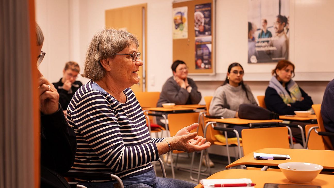 Person mit gestreiftem Pullover spricht in einem Raum, andere Personen sitzen an Tischen, im Hintergrund Plakate an der Wand.