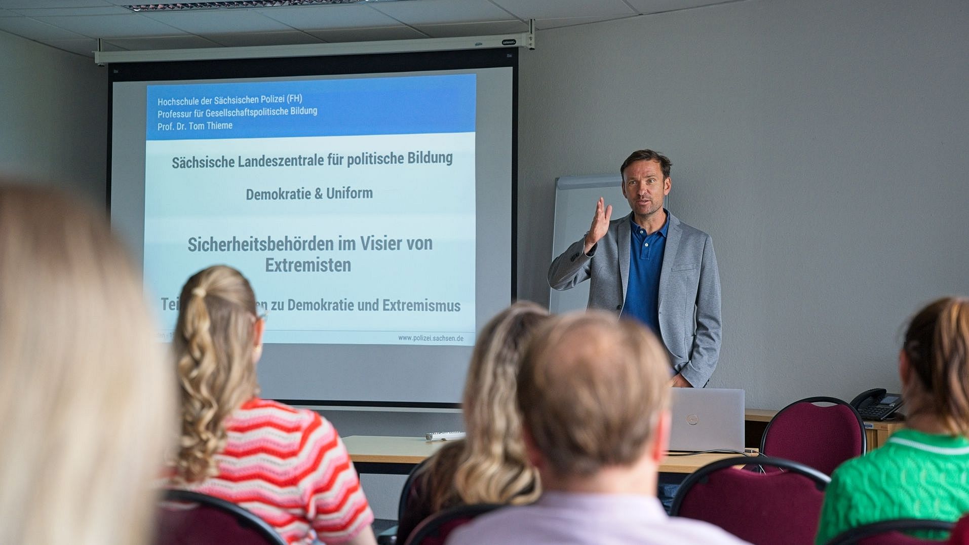 Prof. Dr. Tom Thieme spricht vor Teilnehmenden einer Fortbildung im Hauptzollamt Dresden. Auf der Leinwand ist die Titelfolie der Veranstaltung „Demokratie & Uniform“ zu sehen.