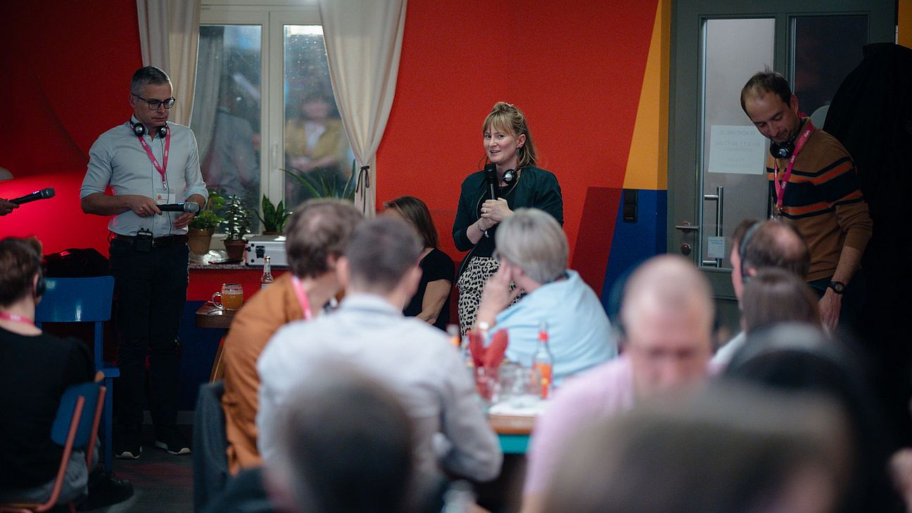 Drei Personen stehen vor einer bunt gestrichenen Wand mit Vorhängen, mehrere Personen sitzen an Tischen im Vordergrund.