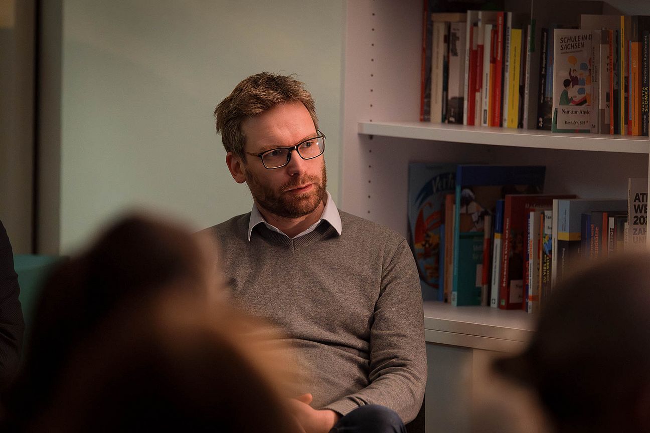 Mann mit Pullover sitzt vor einem Bücherregal in einem Raum mit mehreren Personen im Vordergrund.