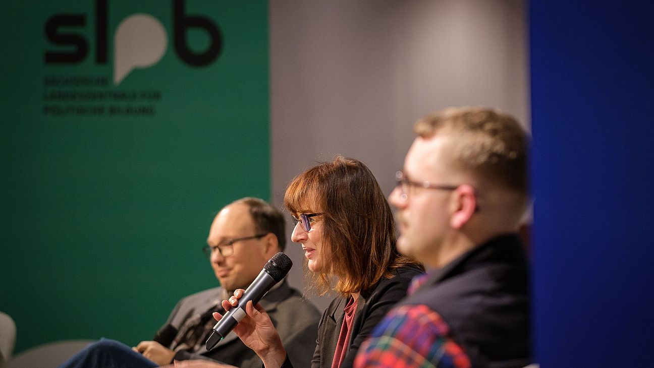 rei Personen sitzen nebeneinander, eine Frau hält ein Mikrofon, im Hintergrund ein grünes Schild mit schwarzem Text und Logo.