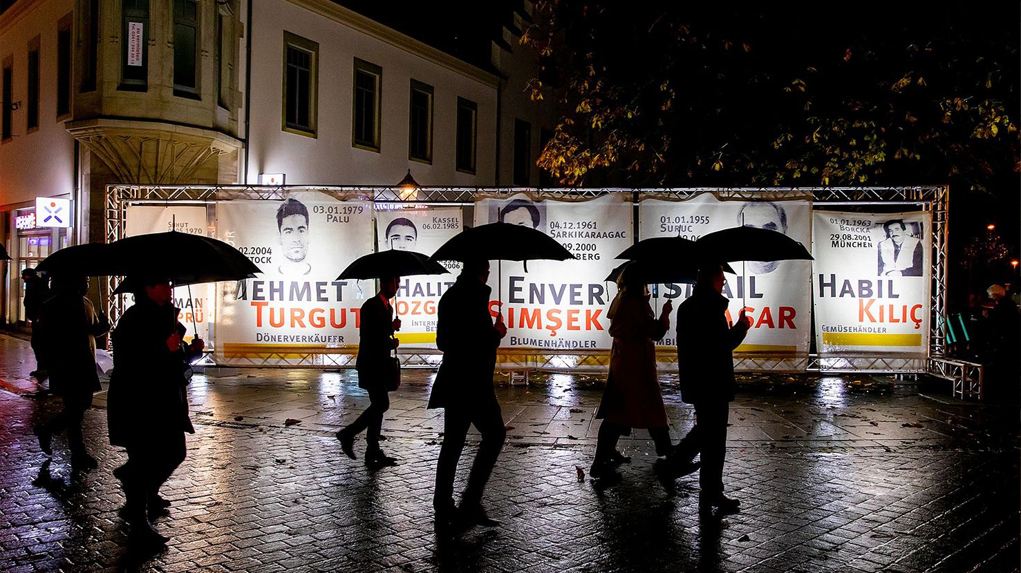 Gedenken an die Opfer der NSU-Morde in der Zwickauer Innenstadt. Menschen laufen abends mit Regenschirmen an Plakate mit Gesichtern und Namen der Opfern entlang.